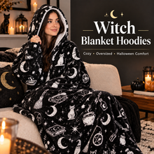 Load image into Gallery viewer, A woman sits on a couch wearing a black hooded blanket hoodie with a white witch-themed print featuring moons, stars, cauldrons, broomsticks, and spells. The scene is styled in a cozy living room with candles, pumpkins, and a dark wall with the text 'Witch Blanket Hoodies' and 'Cozy • Oversized • Halloween Comfort'.