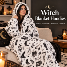 Load image into Gallery viewer, A woman sits on a couch wearing a white hooded blanket hoodie covered in black witch-themed prints including moons, cats, cauldrons, skulls, and the word 'WITCH.' Text on the right reads 'Witch Blanket Hoodies' with smaller copy 'Cozy • Oversized • Halloween Comfort.
